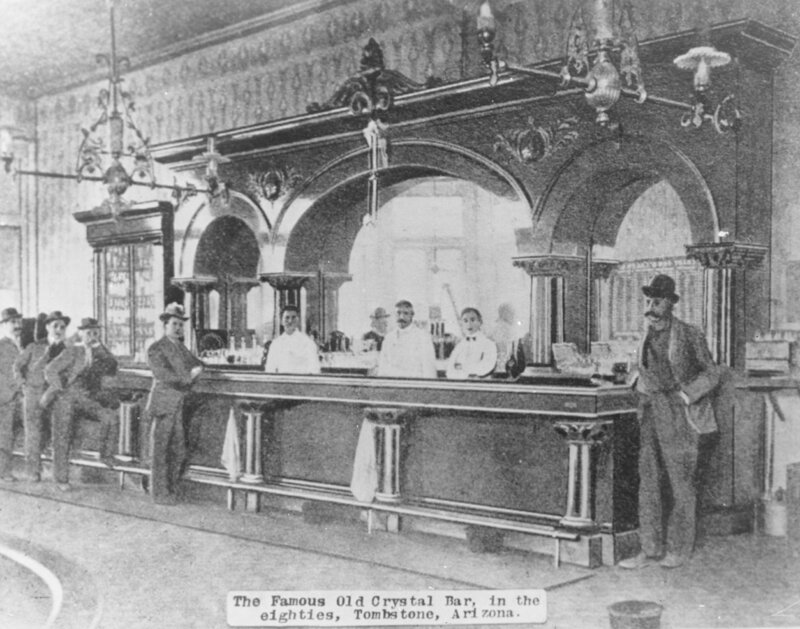 Interior of an ornate saloon with a long carved bar and arched back bar mirrors, three bartenders in white, and customers in suits and hats standing along the bar.